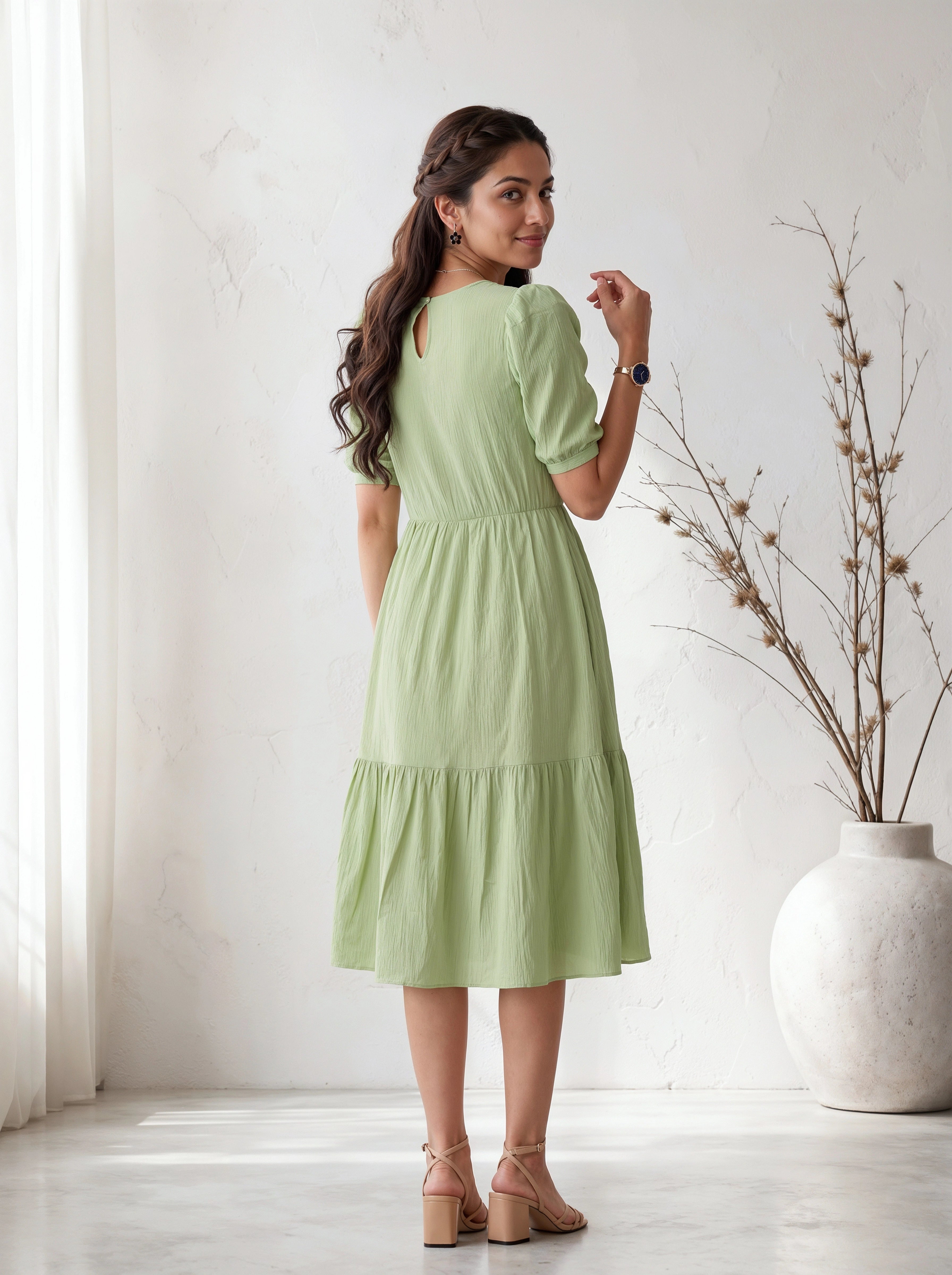 Woman wearing a light green dress standing in a bright room with a vase and plant in the background.