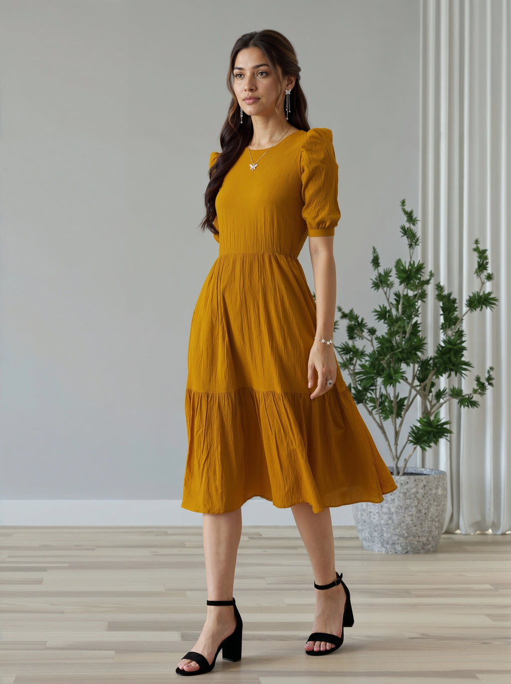 Woman wearing a mustard yellow dress standing in a room with a plant and light-colored walls.
