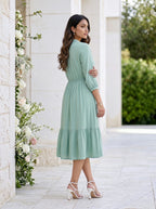 Woman in a light green dress standing in a decorative outdoor setting with flowers and a white wall.