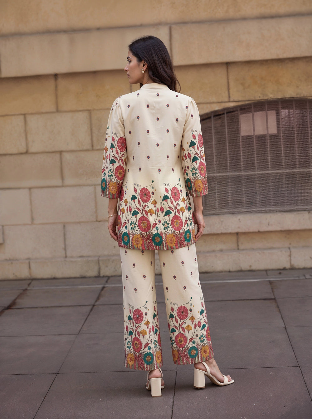 A woman stands with her back to the camera wearing a cream-colored embroidered kurta and matching wide-leg pants. The outfit features colorful floral patterns in red, green, and orange, with small brown polka dots throughout.