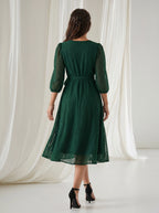 Woman wearing a green lace dress standing in front of white curtains.