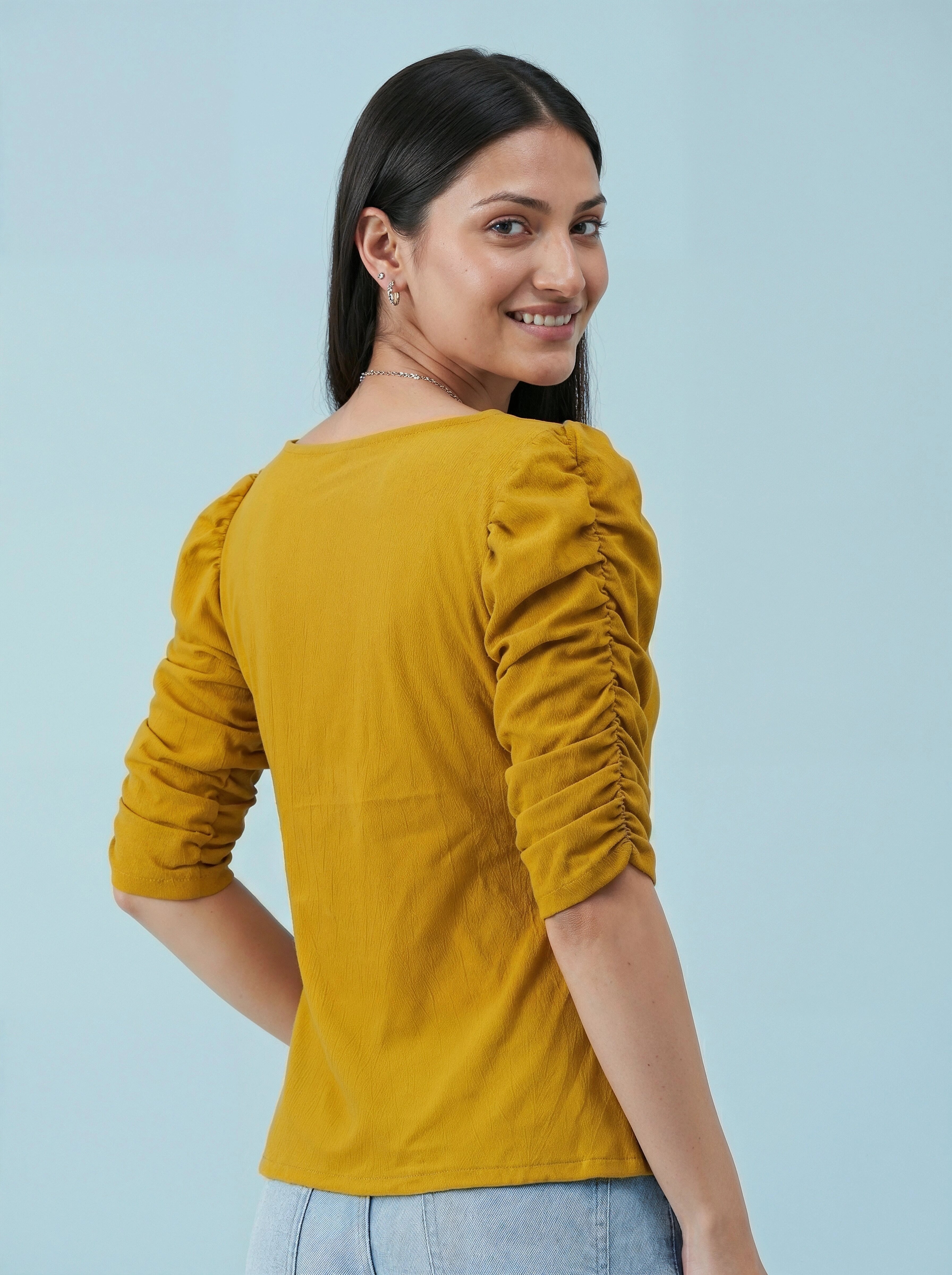 Woman wearing a mustard yellow top with ruched sleeves against a light blue background