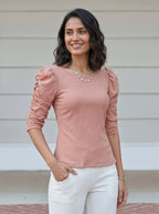 Woman wearing a pink top with ruffled sleeves and white pants against a neutral background