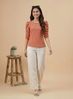 Woman wearing a rust-colored top and white pants standing against a beige background