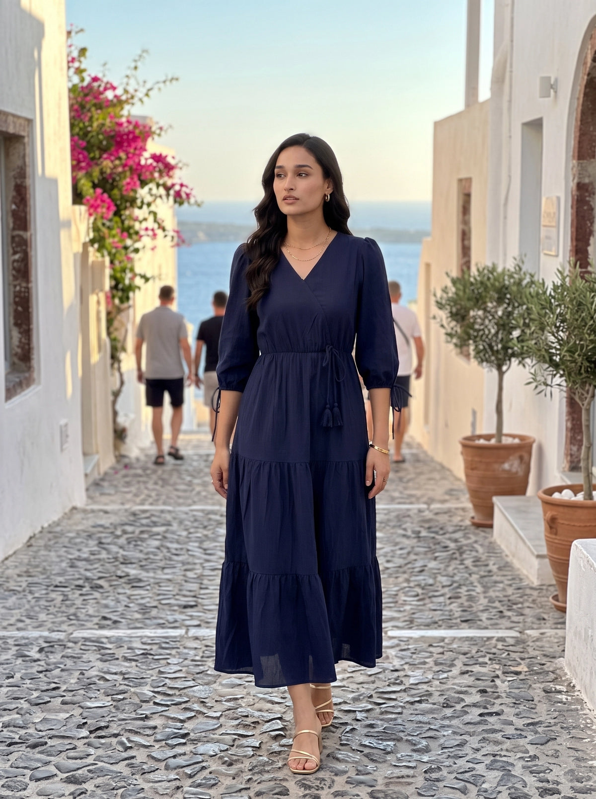 A woman wearing a navy blue midi dress walks down a cobblestone street in a Mediterranean-style village with white buildings and a view of the sea in the background.