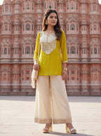 A woman stands outdoors in front of a large pink sandstone palace, wearing a bright yellow kurta with a heavily embellished silver and gold neckline, paired with wide-leg cream pants and holding a cream dupatta.