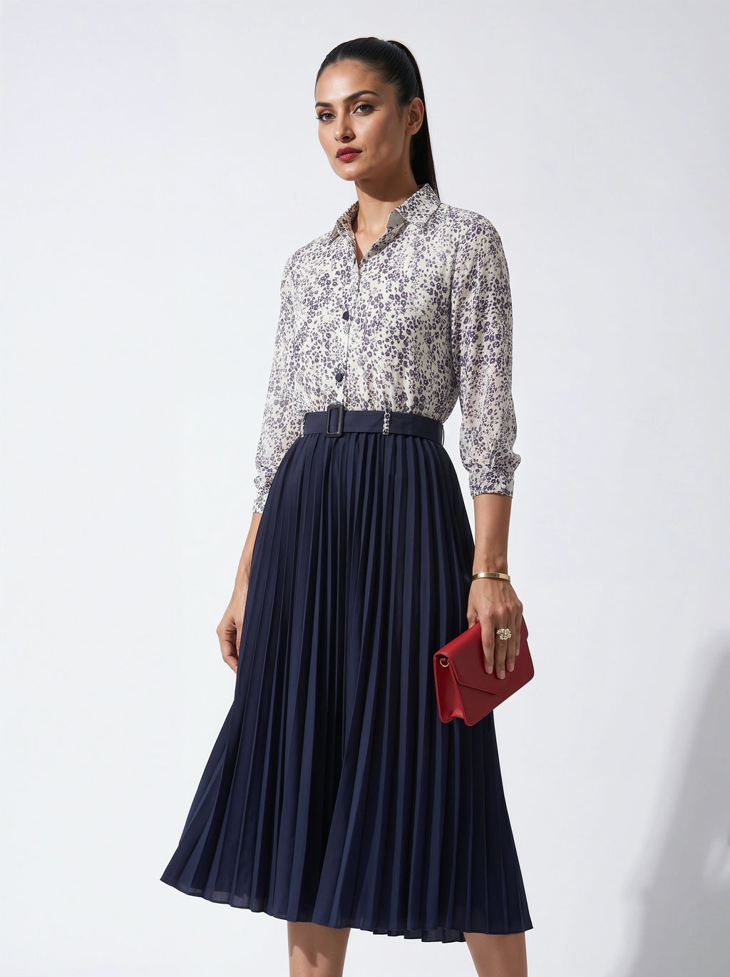 Woman wearing a floral Shirt and navy pleated skirt with a red clutch against a white background
