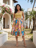 Woman wearing a yellow top and colorful floral skirt standing outdoors with a building and palm trees in the background.