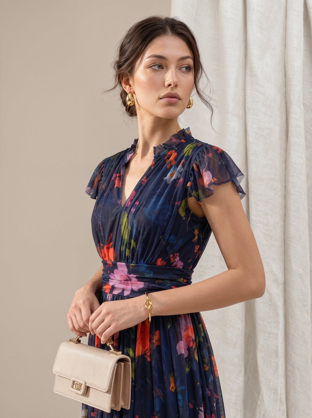 A woman wearing a navy blue floral chiffon dress with ruffled sleeves and a tie waist, holding a beige handbag. She is posed against a neutral beige background with a draped white curtain.