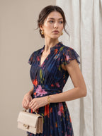 A woman wearing a navy blue floral chiffon dress with ruffled sleeves and a tie waist, holding a beige handbag. She is posed against a neutral beige background with a draped white curtain.