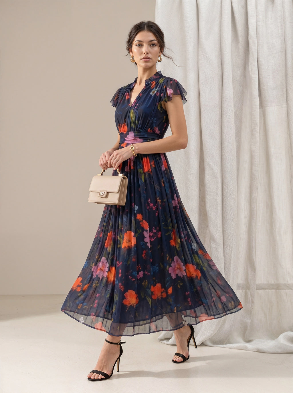 A woman models a navy blue midi dress with a vibrant orange, pink, and red floral print. She is holding a beige handbag and wearing black high-heeled sandals, standing in front of a neutral beige backdrop with white draped curtains.