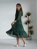 Woman wearing a green dress standing in a minimalistic room with a plant.