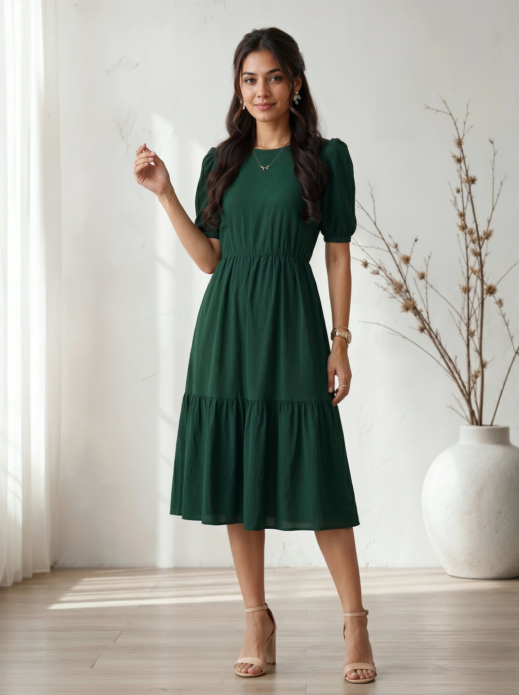 Woman wearing a green dress standing in a room with a white curtain and vase.