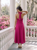 A woman wearing a sage green tiered dress with three-quarter puff sleeves and an asymmetrical draped neckline, shown in a warm indoor setting.