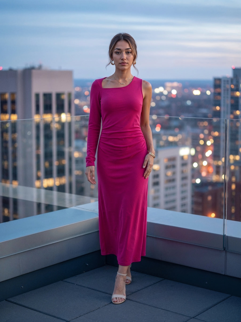 A woman stands on a rooftop terrace at dusk wearing a bright fuchsia pink long-sleeve midi dress with a one-shoulder neckline and gathered waist detail. The background shows a blurred city skyline with illuminated windows.