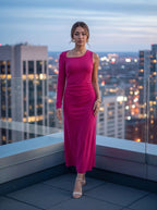 A woman stands on a rooftop terrace at dusk wearing a bright fuchsia pink long-sleeve midi dress with a one-shoulder neckline and gathered waist detail. The background shows a blurred city skyline with illuminated windows.