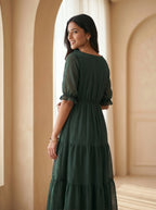 Woman wearing a green dress standing in a room with beige walls and a white curtain.