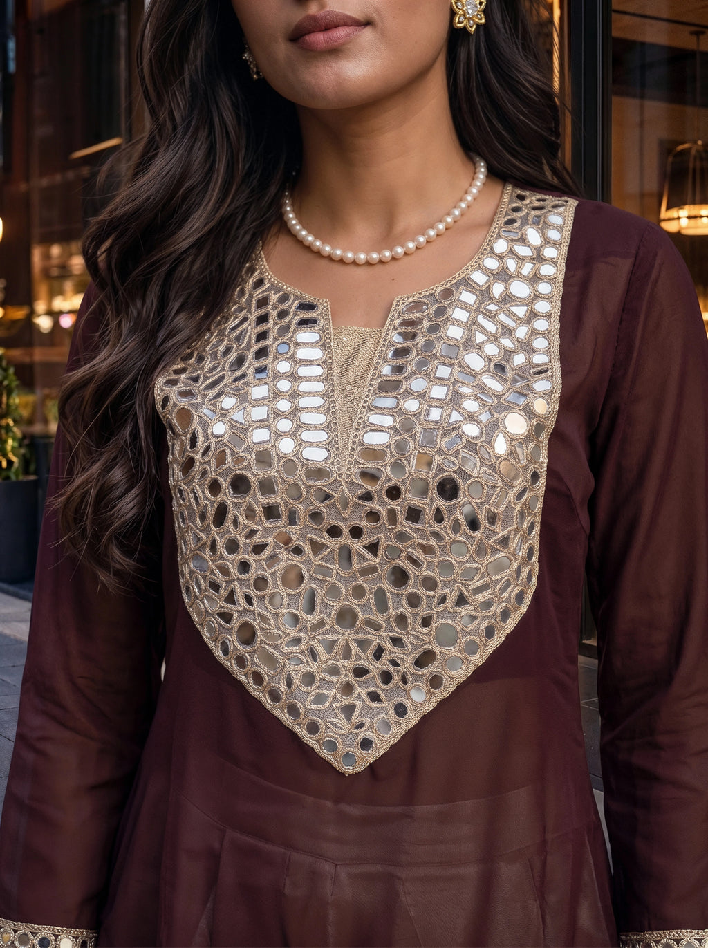 A close-up of a woman wearing a deep maroon long kurta with an intricate beige and silver mirror-work embroidered yoke on the chest, accessorized with a pearl necklace.