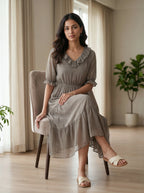 Woman in a gray dress sitting on a chair in a room with plants and curtains.