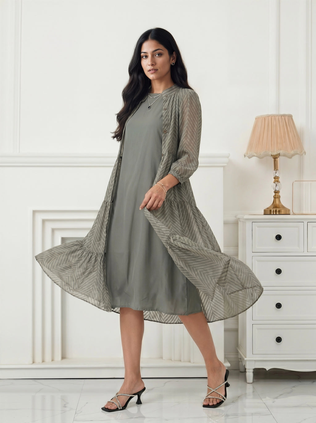 Woman wearing a long green dress with a patterned overlay in a room with white walls and a lamp.