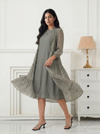 Woman wearing a long green dress with a patterned overlay in a room with white walls and a lamp.