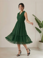Woman wearing a green dress standing indoors with a plant in the background