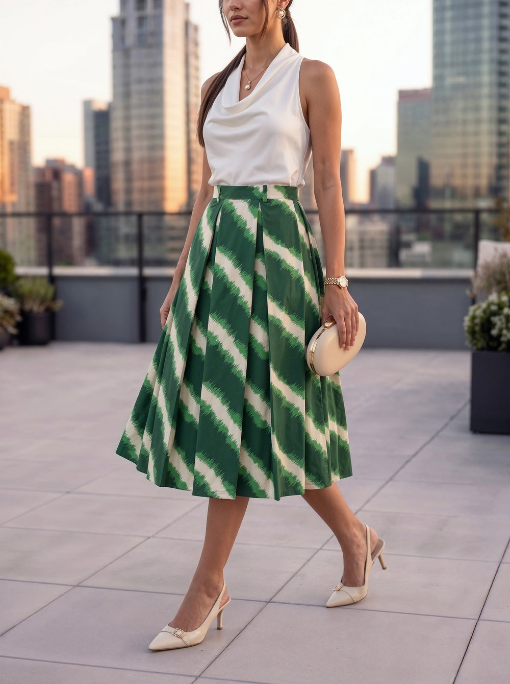 A woman models a sleeveless white top and a pleated midi skirt with a green and white diagonal tie-dye pattern, styled with cream heels and a small clutch, set against a city skyline at sunset.