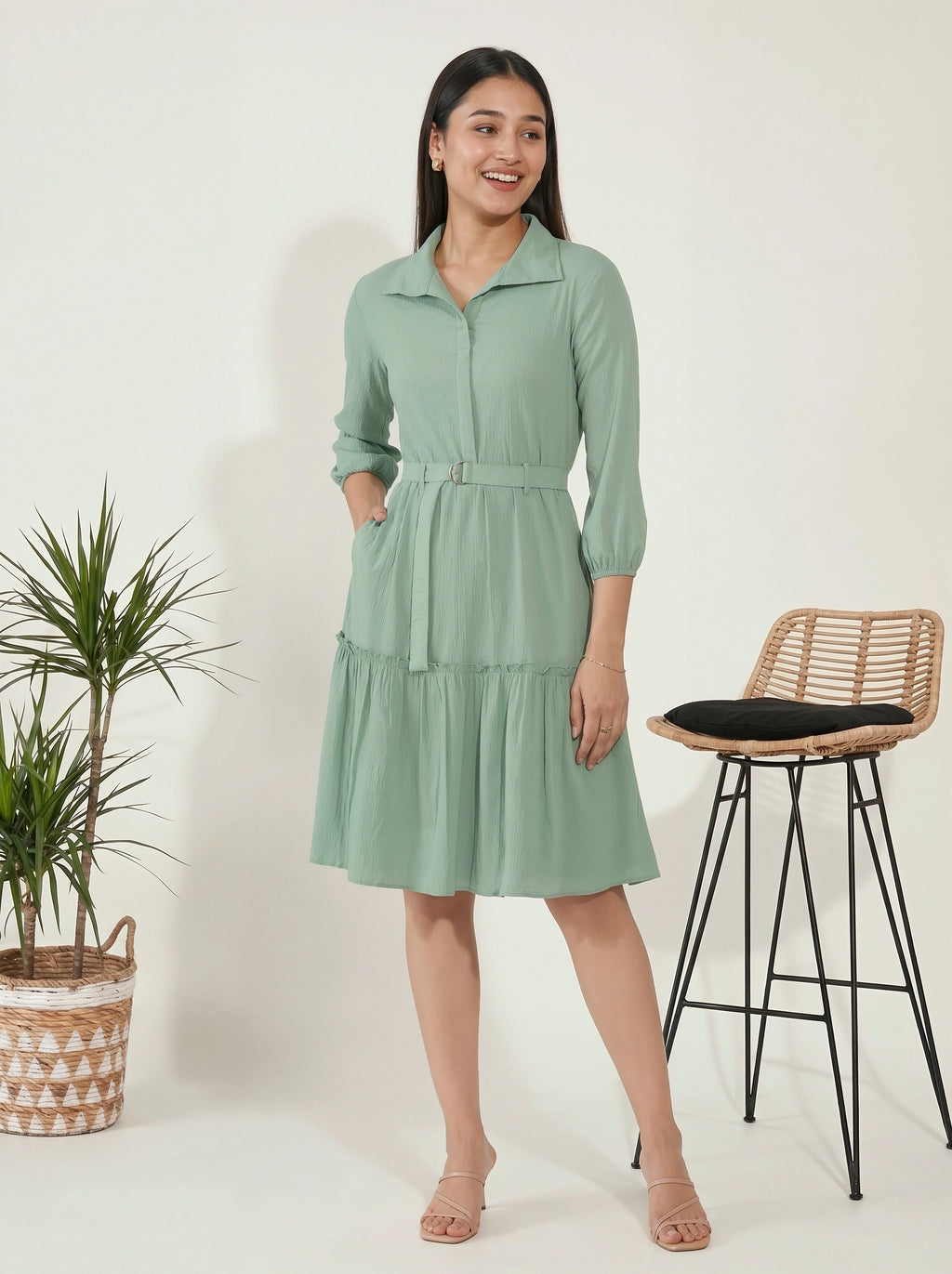 Woman wearing a light green dress standing next to a plant and a stool on a white background