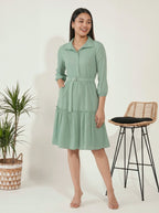 Woman wearing a light green dress standing next to a plant and a stool on a white background
