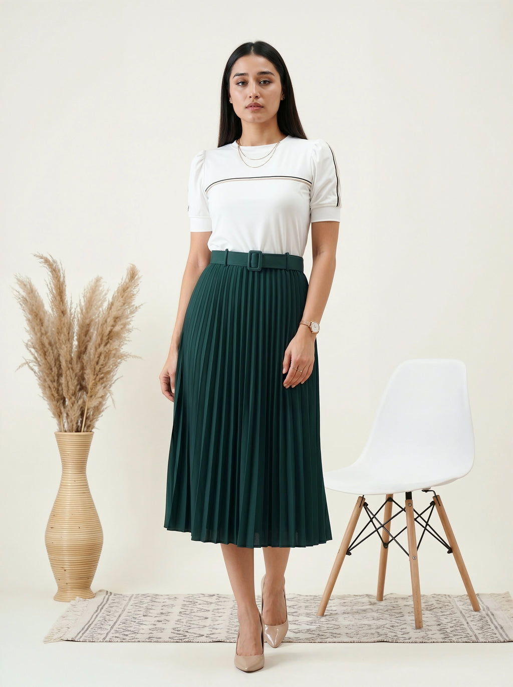Woman wearing a white top and green pleated skirt standing in a minimalistic room with a chair and vase.