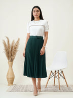 Woman wearing a white top and green pleated skirt standing in a minimalistic room with a chair and vase.