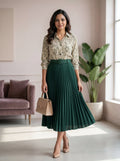 Woman in a green pleated skirt and patterned blouse standing in a bright room with a plant and sofa.