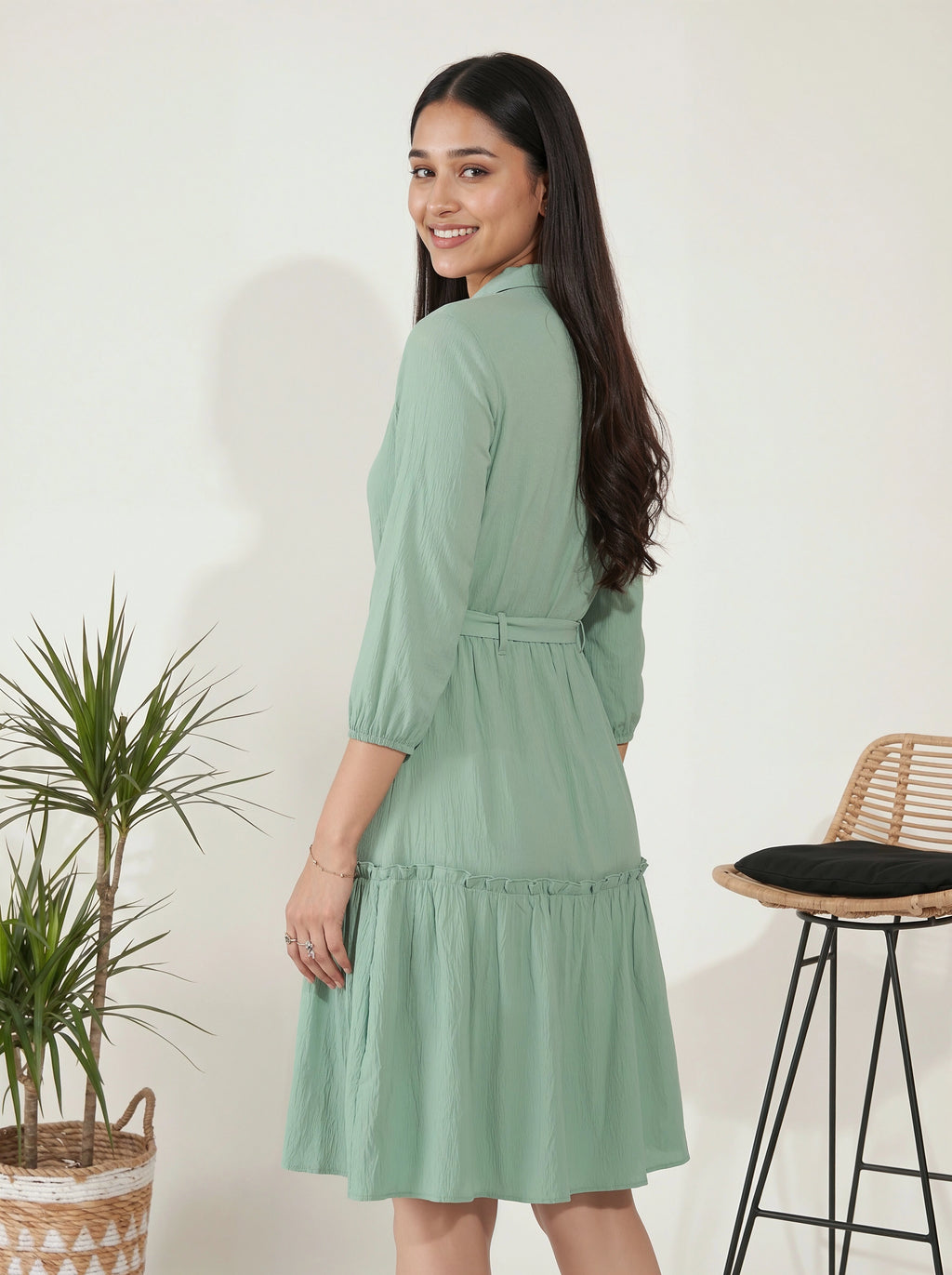 Woman wearing a green dress standing next to a plant and stool indoors.
