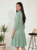 Woman wearing a green dress standing next to a plant and stool indoors.