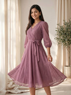 Woman wearing a purple dress standing in a softly lit room with curtains and a plant.