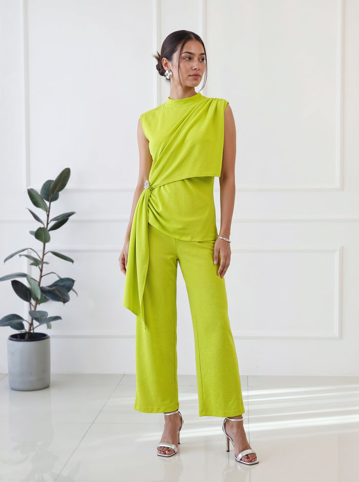A woman models a bright lime green one-piece jumpsuit with a draped asymmetrical neckline, side tie detail, and wide-leg cropped pants, standing in a bright white room with a potted plant.