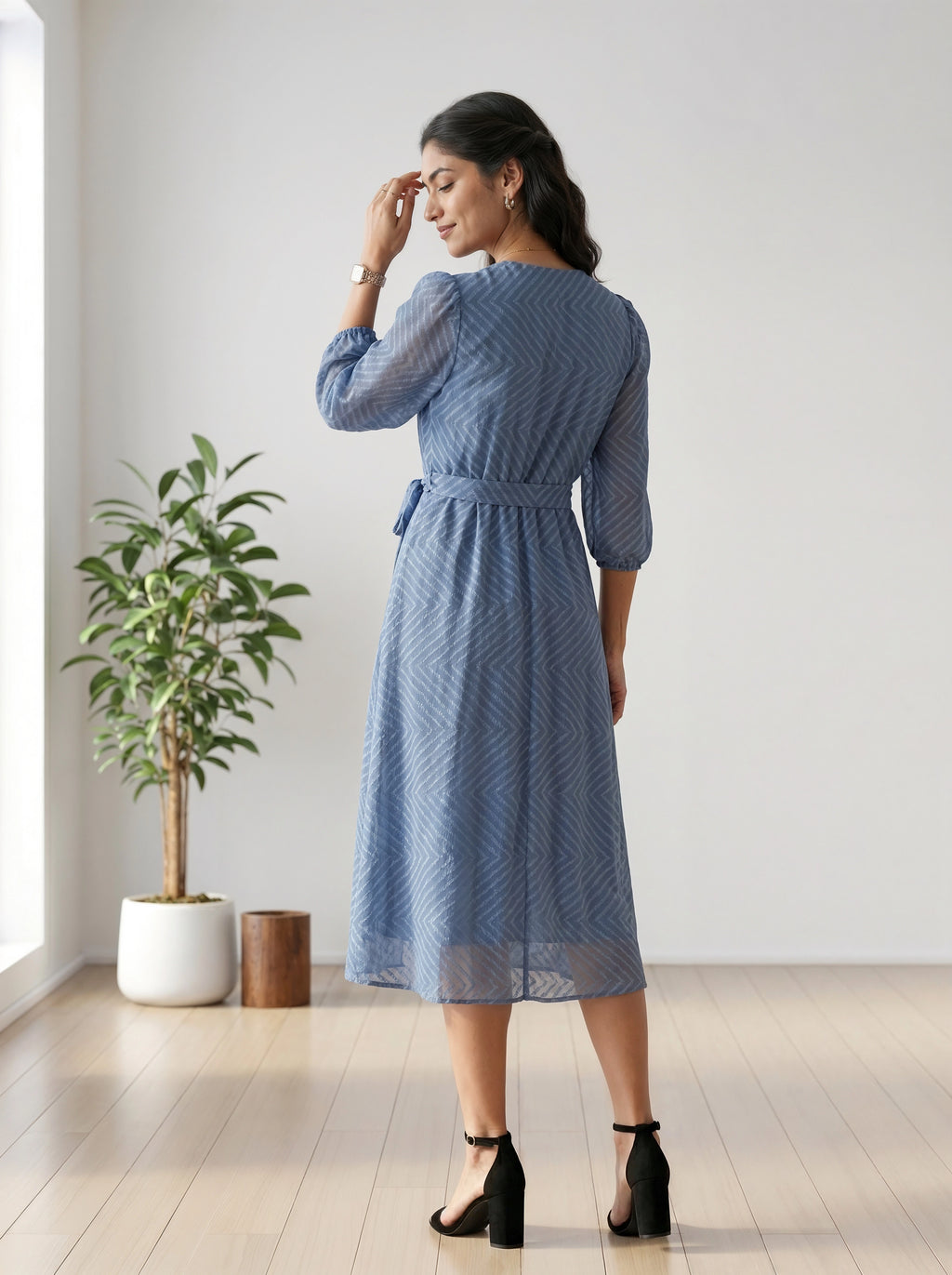 Woman wearing a blue dress standing in a room with a plant and wooden stool.