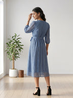 Woman wearing a blue dress standing in a room with a plant and wooden stool.