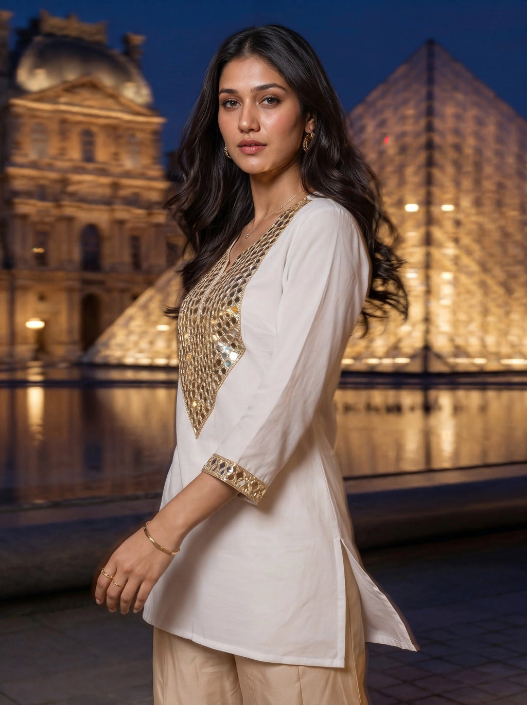A woman modeling a white kurta with gold sequin embellishments on the neckline and sleeve cuffs, paired with beige trousers, set against a nighttime background of the Louvre Museum and the Louvre Pyramid.