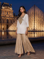 A woman models a white and gold embroidered tunic with wide-leg beige trousers, standing in front of the illuminated Louvre Museum and glass pyramid at night.