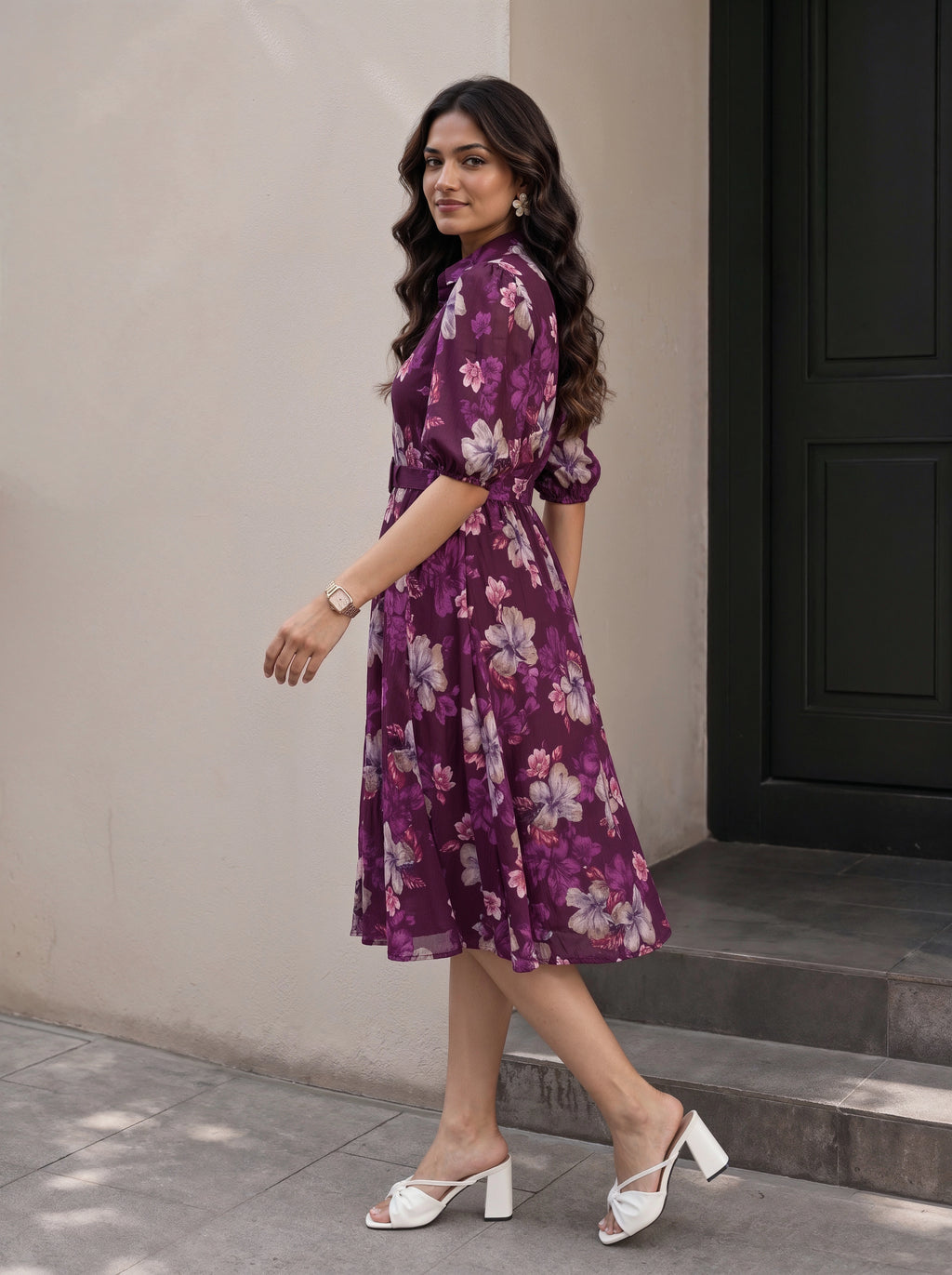 A woman modeling a black midi dress with three-quarter sleeves, a high neckline with a keyhole cutout, and a tiered ruffle hem, set against a warm beige studio background with a small wooden stool and plant.