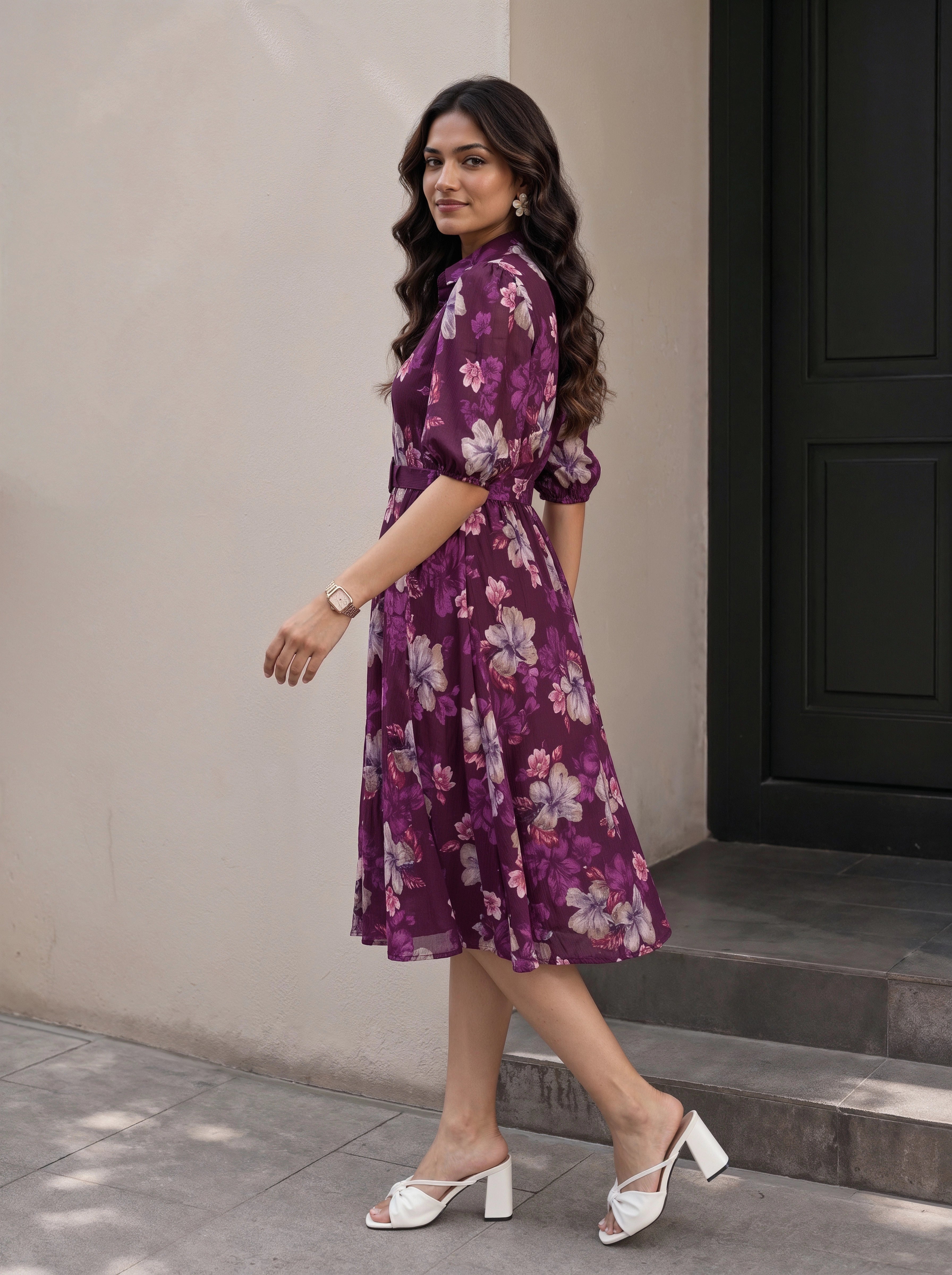 A woman modeling a black midi dress with three-quarter sleeves, a high neckline with a keyhole cutout, and a tiered ruffle hem, set against a warm beige studio background with a small wooden stool and plant.