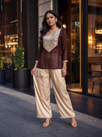 A woman models a two-piece traditional outfit consisting of a dark brown long-sleeve kurta with a metallic gold embroidered yoke, paired with wide-leg champagne satin pants, standing on a city sidewalk.