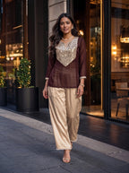 A woman models a two-piece traditional outfit consisting of a brown long-sleeve kurta with a heavily embellished gold sequin V-neck bodice, paired with wide-leg beige satin pants with a beaded hem, set against a modern building entrance at dusk.