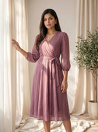 Woman wearing a pink dress standing in a softly lit room with curtains and a plant.