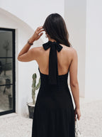 A woman shown from behind wearing a black halter-neck maxi dress with a large bow detail at the back, standing in a bright, modern white architectural setting.