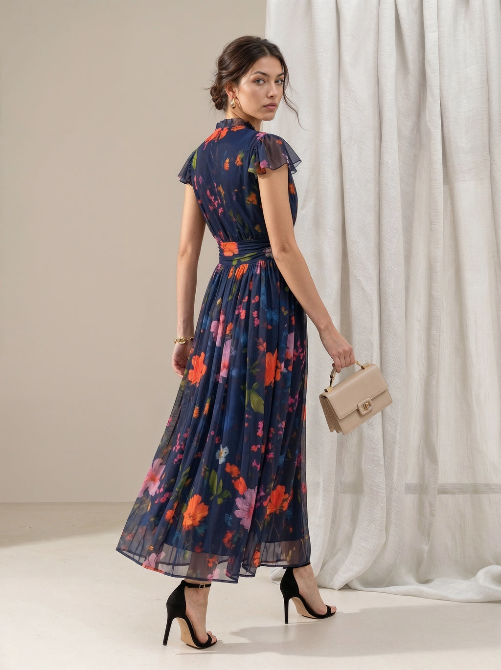 A woman models a navy blue maxi dress with a vibrant multicolor floral print, featuring ruffled cap sleeves and a tie-neck detail. She is holding a beige handbag and wearing black high heels, posed in front of a neutral studio backdrop with draped white fabric.