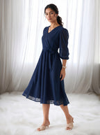Woman wearing a navy blue dress with sheer sleeves against a white curtain background