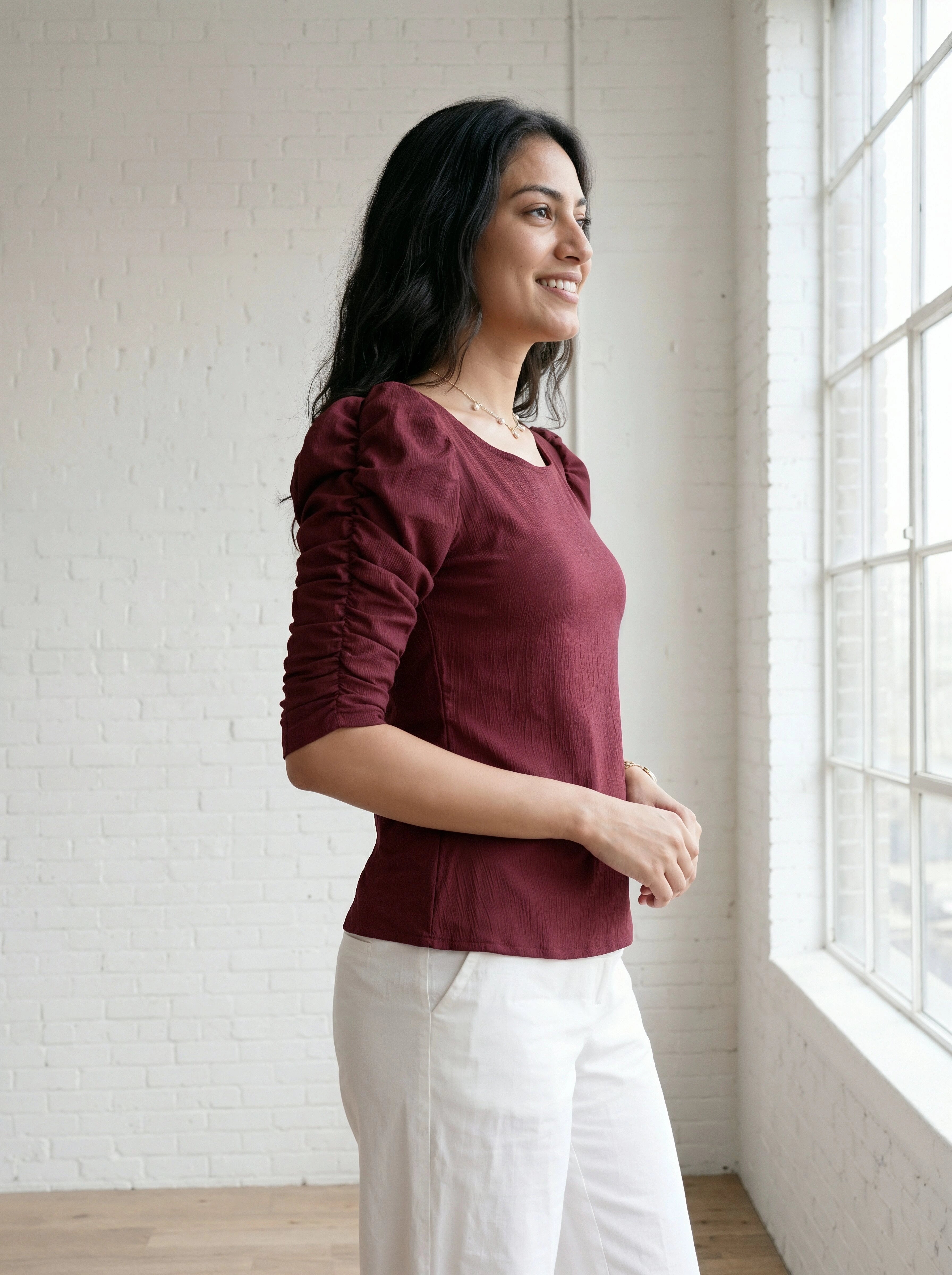Woman wearing a burgundy top and white pants standing in a room with a white brick wall and large window.