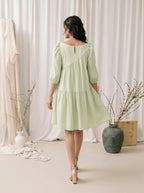 Woman in a light green dress standing in a room with white curtains and decorative vases.